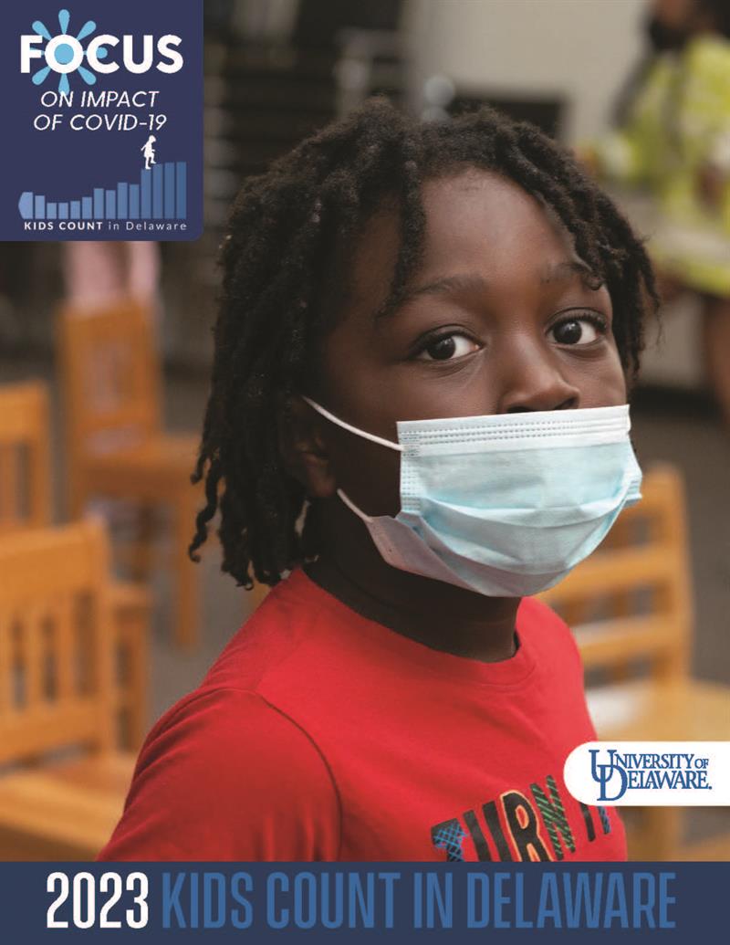 KIDS COUNT in Delaware 2023 FOCUS on Impact of COVID-19 book cover: image of masked boy wearing red shirt standing in a classroom surrounded by wooden chairs
