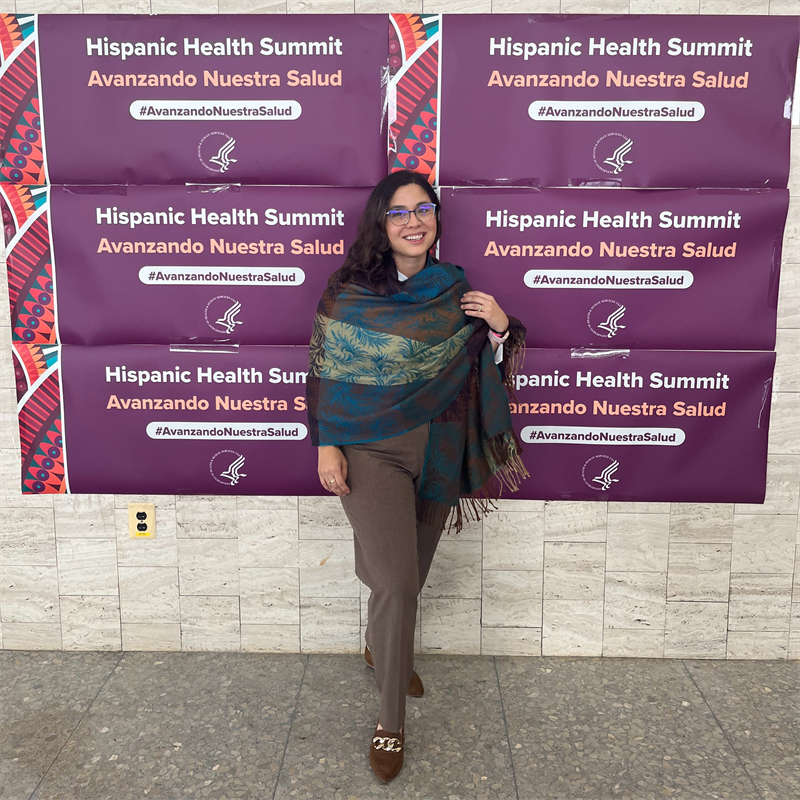 Dr. Mitsdarffer at the HHS Health Summit standing in front of signage for the event in both English and Spanish.