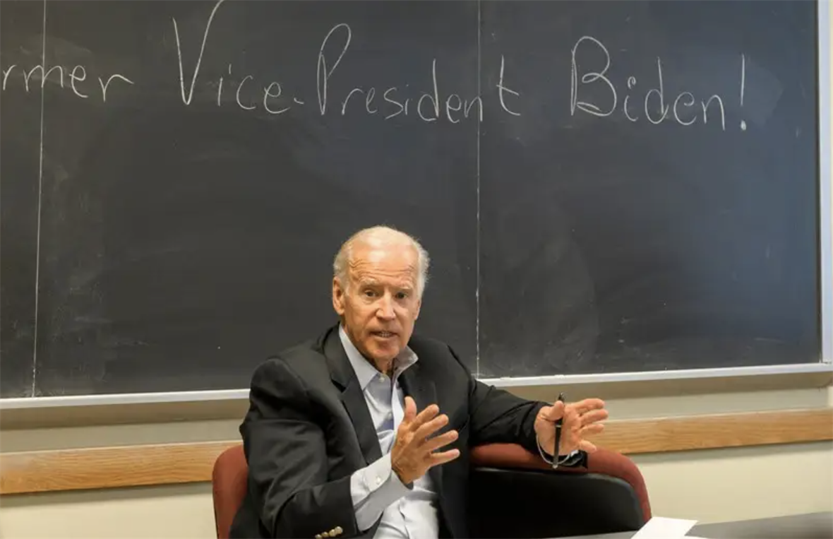 Joe Biden seated and speaking in front of a chalkboard.