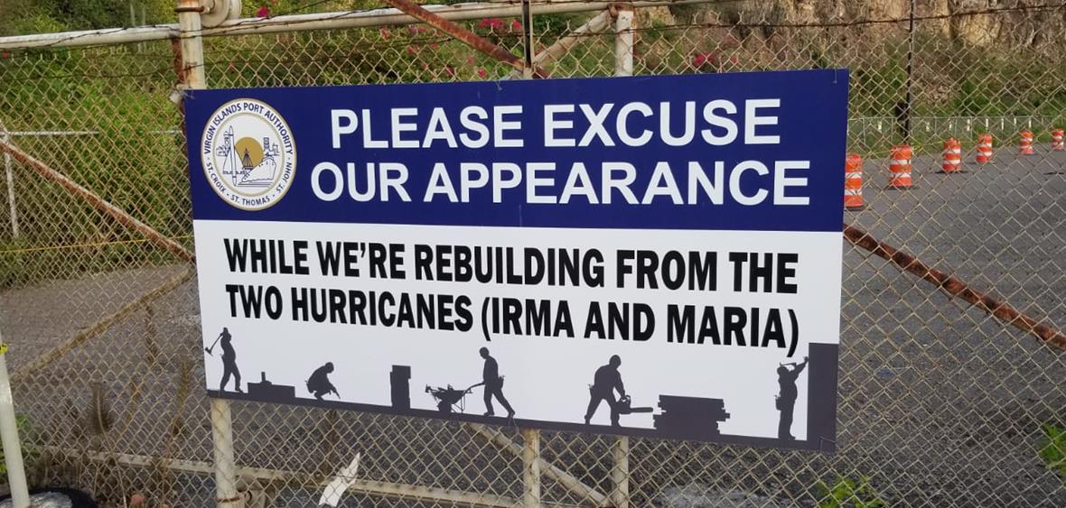 A sign against a fence reads "please excuse our appearance. We are rebuilding from the 2 hurricanes (Irma and Maria)."