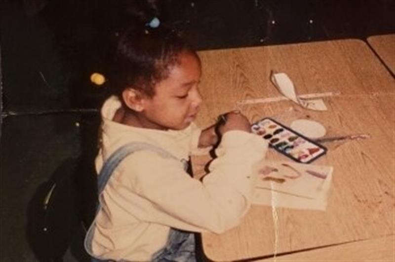 Five-year old sitting at a school desk with watercolor paints,wearing a long sleaved shirt and overalls. 