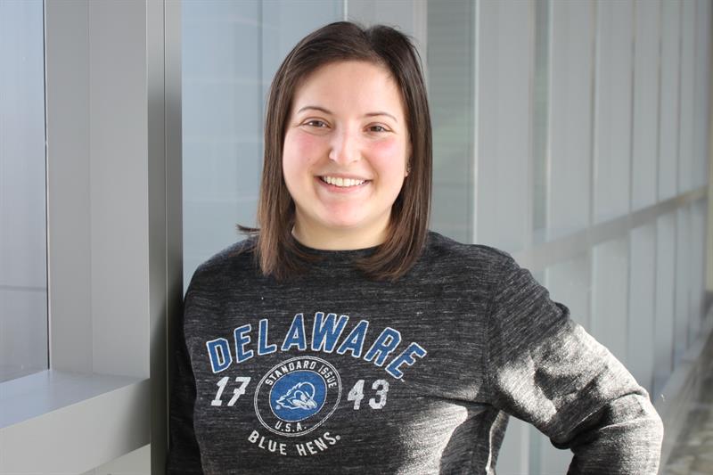 Ellen Schenk wearing a University of Delaware long-sleeved t-shirt, standing against a bay of windows.