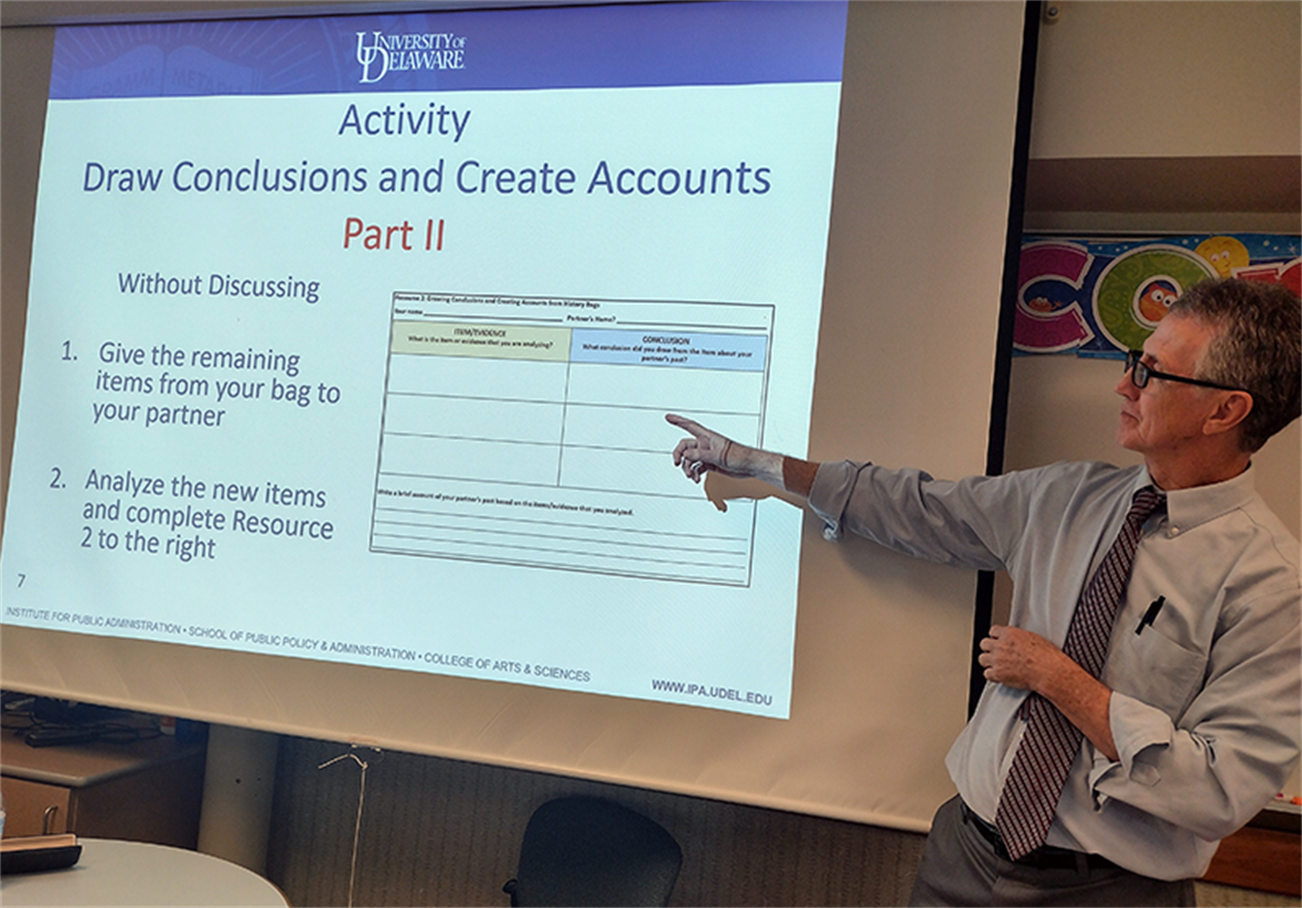 A slide says "Activity: Draw Conclusions and Create Accounts Part II" with the prompts "Give the remaining items from your bag to your partner" and "Analyze the new items and complete Resource 2 to the right". Fran O'Malley is standing next to the projecter screen pointing to the directions.