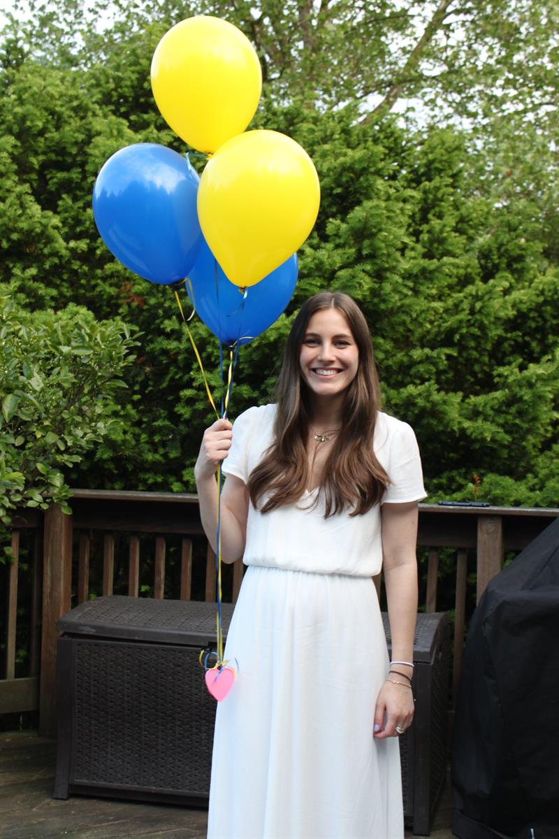 Haley Qaissaunee stands outside in a white dress while holding blue and gold balloons.