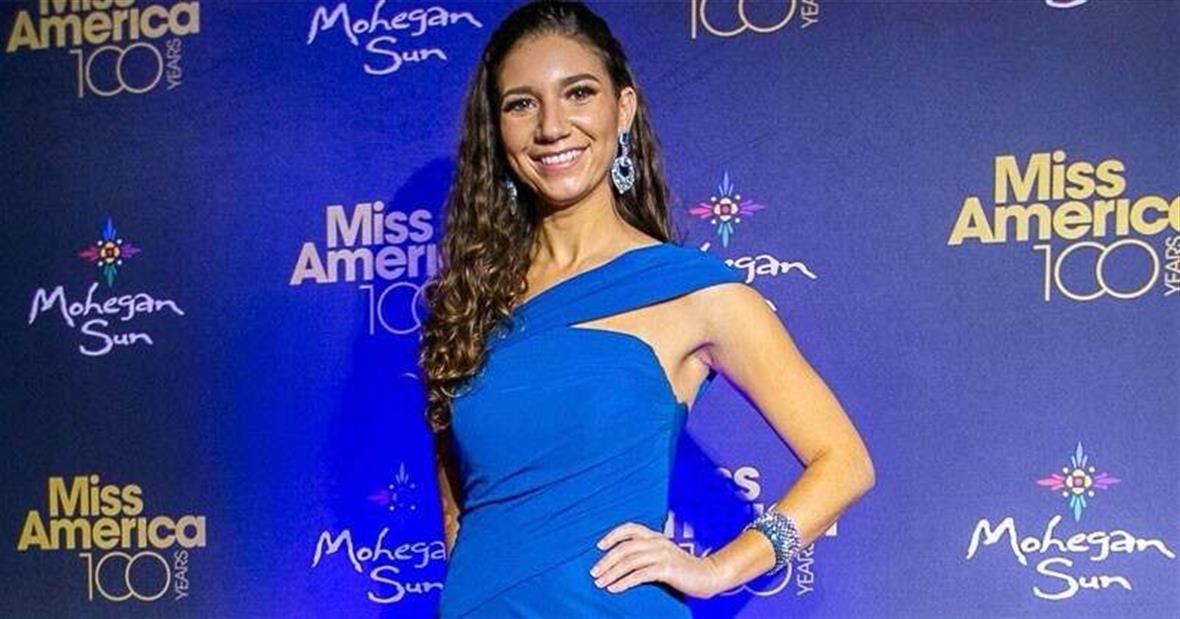 Sophie Phillips poses for a photo in front of the Miss Amereica 100 Years Banner wearing a blue dress and sparkling jewlery. 