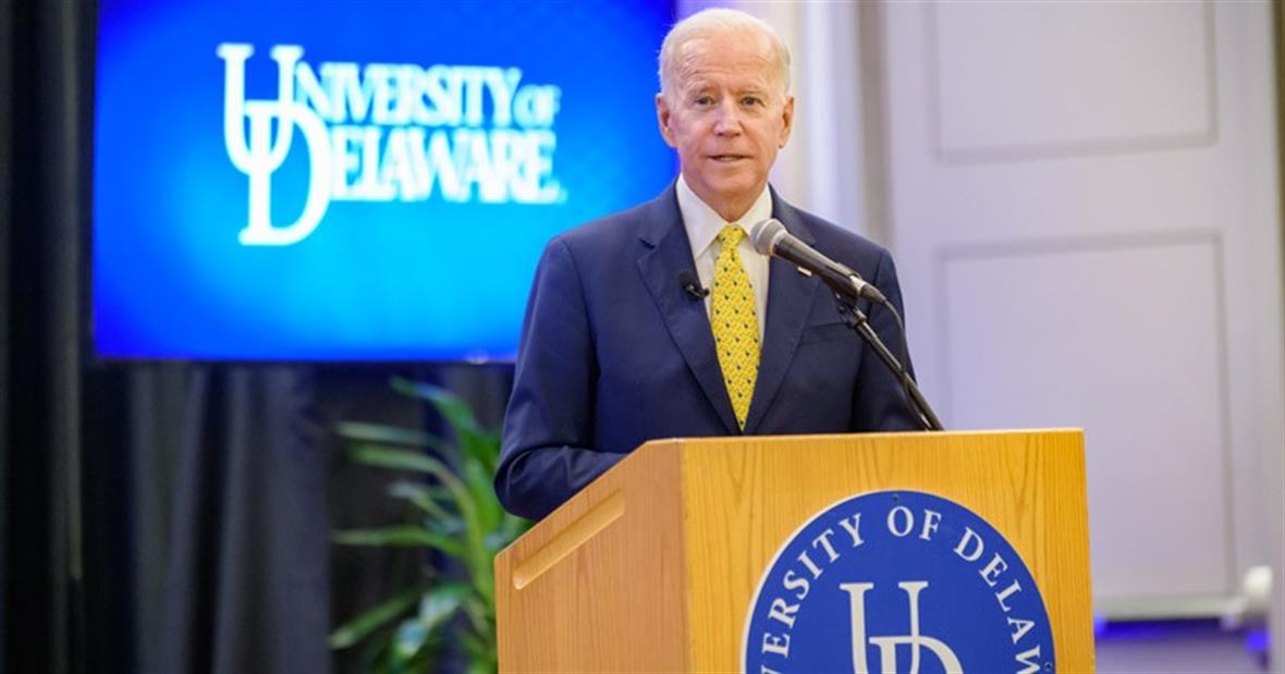 Joe Biden speaking at a podium in front of a blue screen showing the University of Delaware logo.