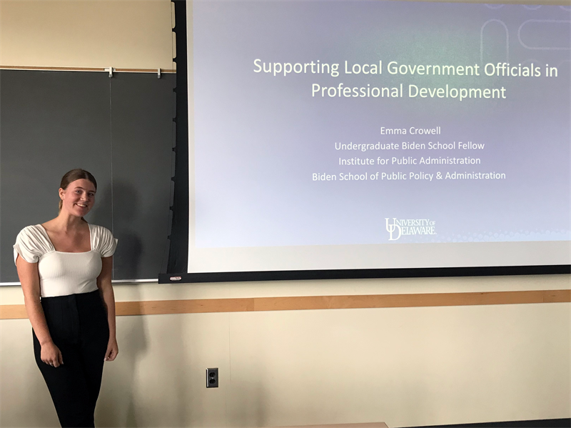 Emma Crowell stands next to the projector screen that displays the title slide of her presentation, "Supporting Local Government Officials in Professional Development."